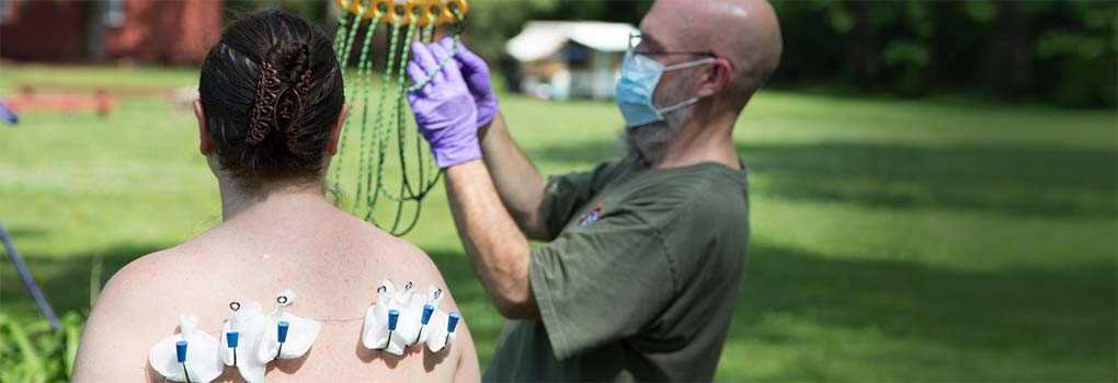 Bald man in surgical mask prepares the rigging, while in the foreground a woman with hooks in her back waits to go up.