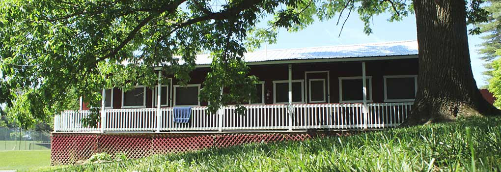 A red painted camping cabin in a greeen field, shaded bya large tree, on a sunny day