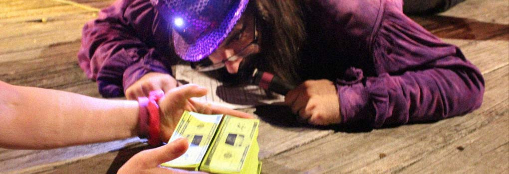 Bo gets down on the stage to closely examine 2 stacks of play money, while wearing a sparkly purple hat.