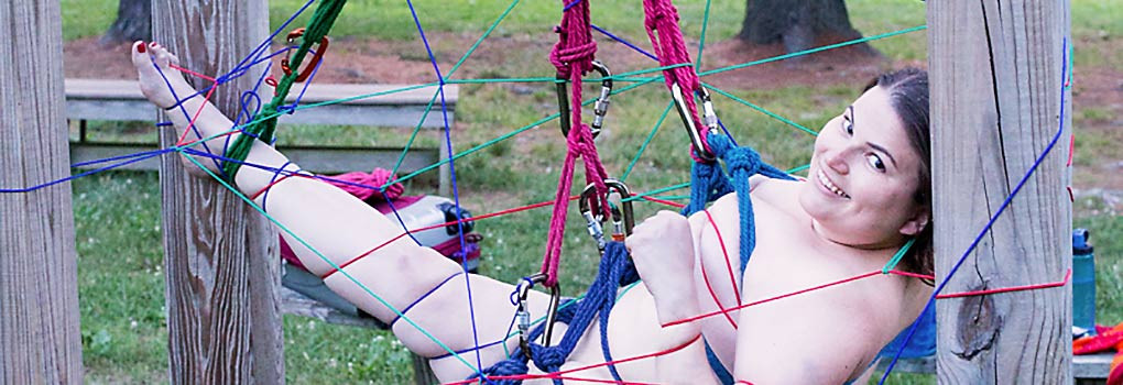 A dark haired smiling girl is suspended outdoors, in blue and red rope