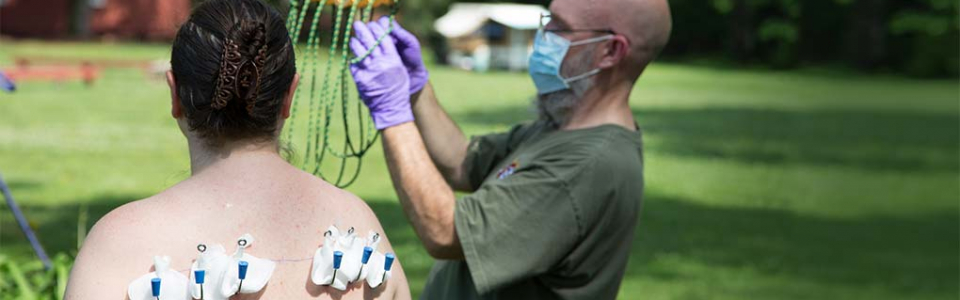 Bald man in surgical mask prepares the rigging, while in the foreground a woman with hooks in her back waits to go up.