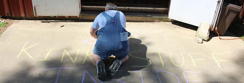 Older male age-player in overalls, writing "kinky things inside!" on the sidewalk in front of a building