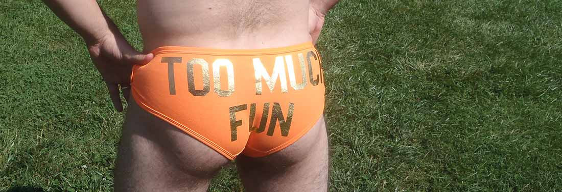 back view of a slender man in tight orange briefs, with "too much fun" in gold letters across the butt